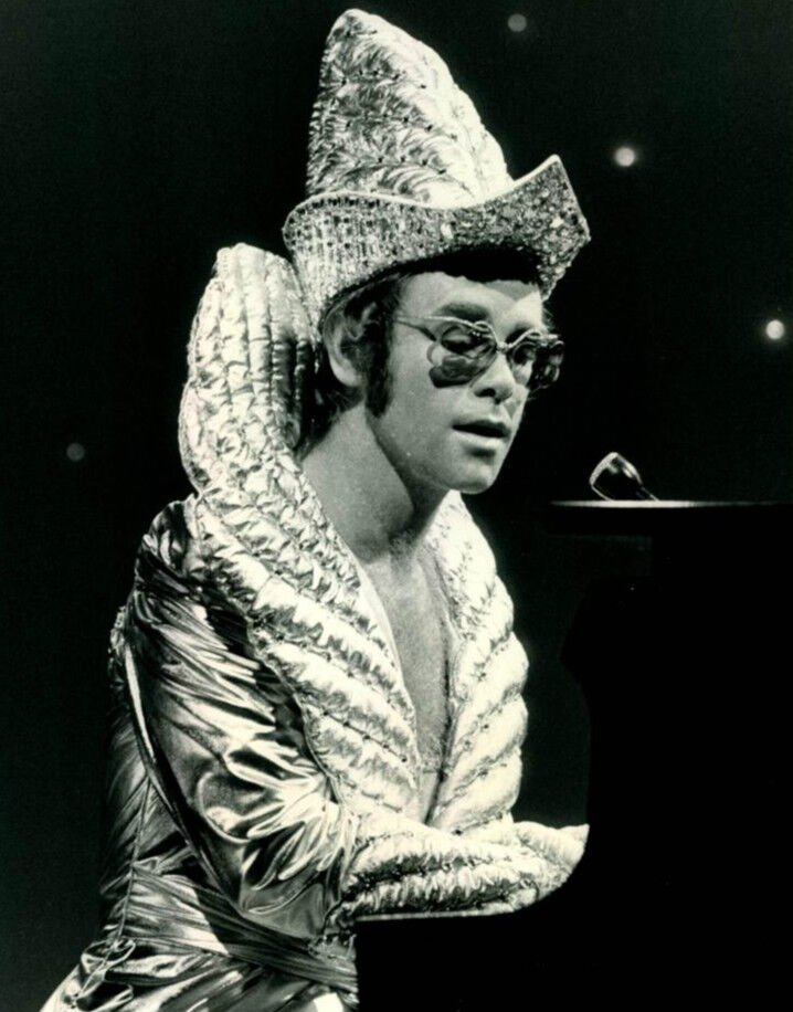 Elton John in a silver quilted costume and tall jeweled hat at a piano, in a 1975 publicity photo for The Cher Show