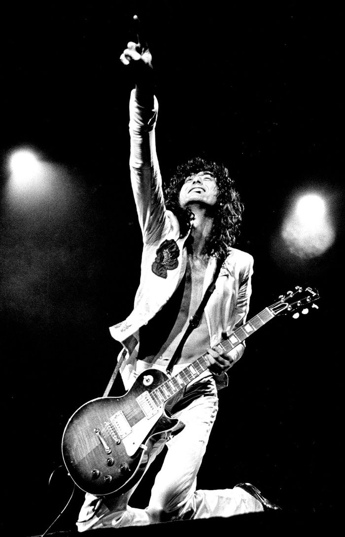 Jimmy Page on one knee with a Gibson Les Paul guitar, one arm raised toward the ceiling, performing live in 1977