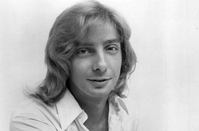Barry Manilow in a black and white 1975 press photo, shoulder-length hair and an open-collared shirt, the face behind the Barry Manilow songs of the 70s